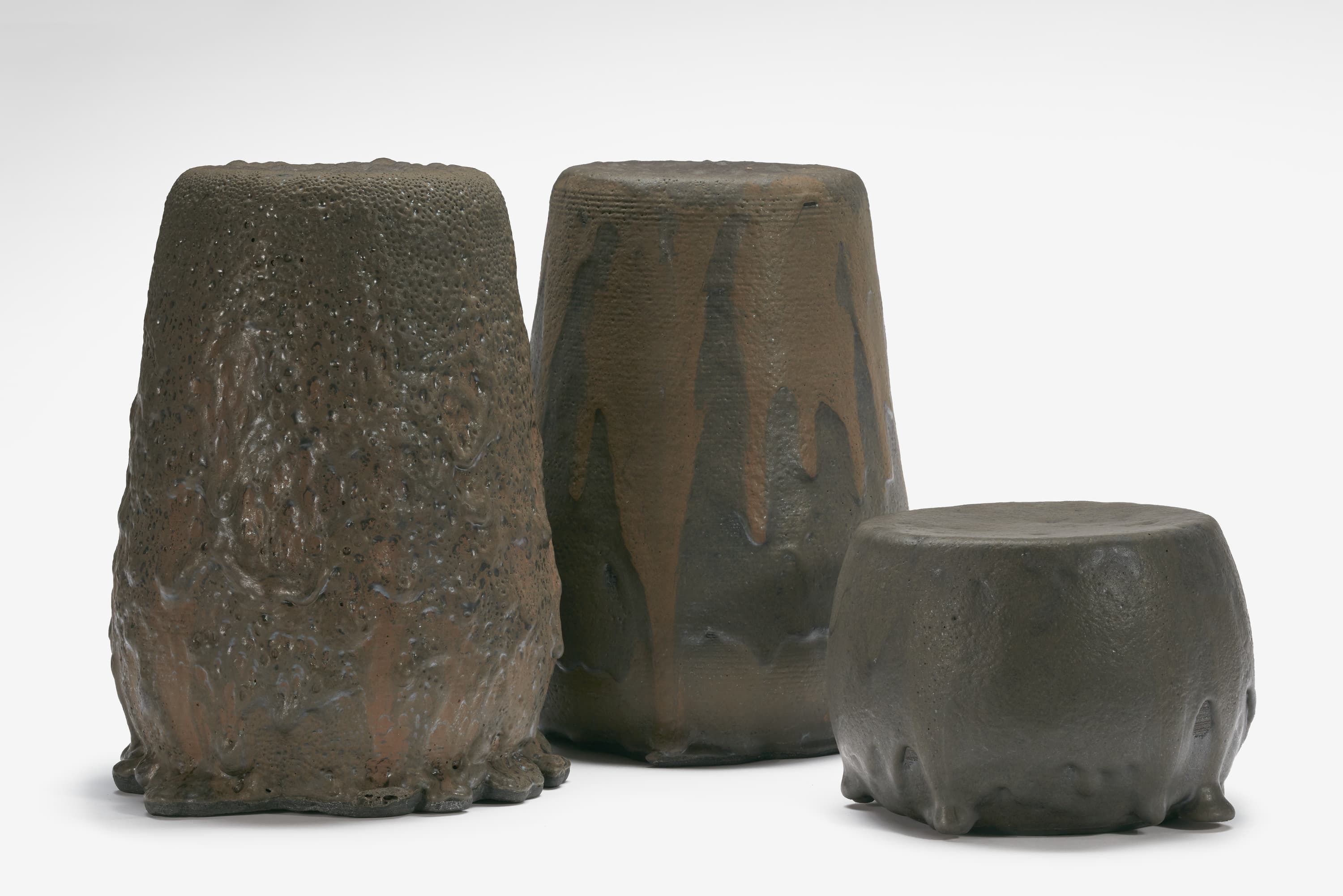 Three cylindrically shaped stools. The left and center stools are the same height; the stool on the right is half the height of the others. The surface of the stools is a dark, brownish gray volcanic lava that drips down the sides.