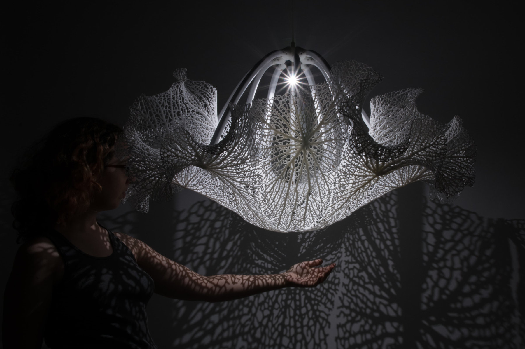 A person stands under Floraform Chandelier and holds out an arm, which is cast in shadows from the artwork, as is the wall behind.