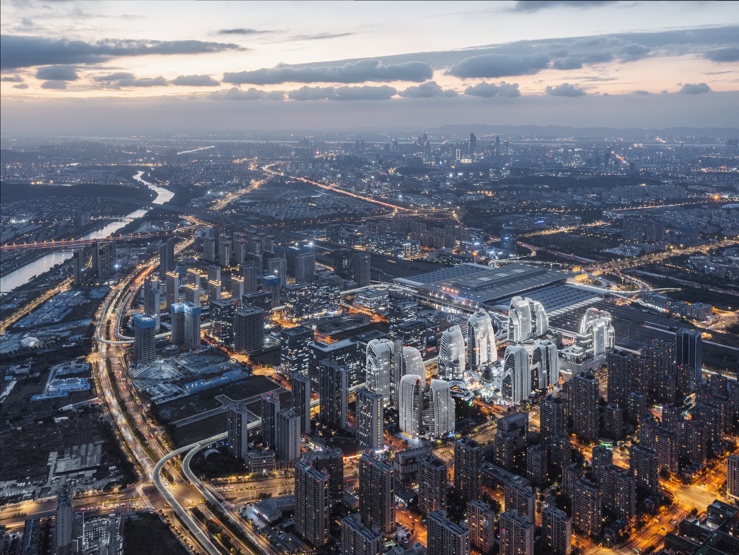 Aerial view at dusk of Nanjing Zendai Himalayas Center. The center is brightly lit and surrounded by the rest of the city; a major road, and a river to the left. Soft gray clouds in the orange sky at the top of the frame.