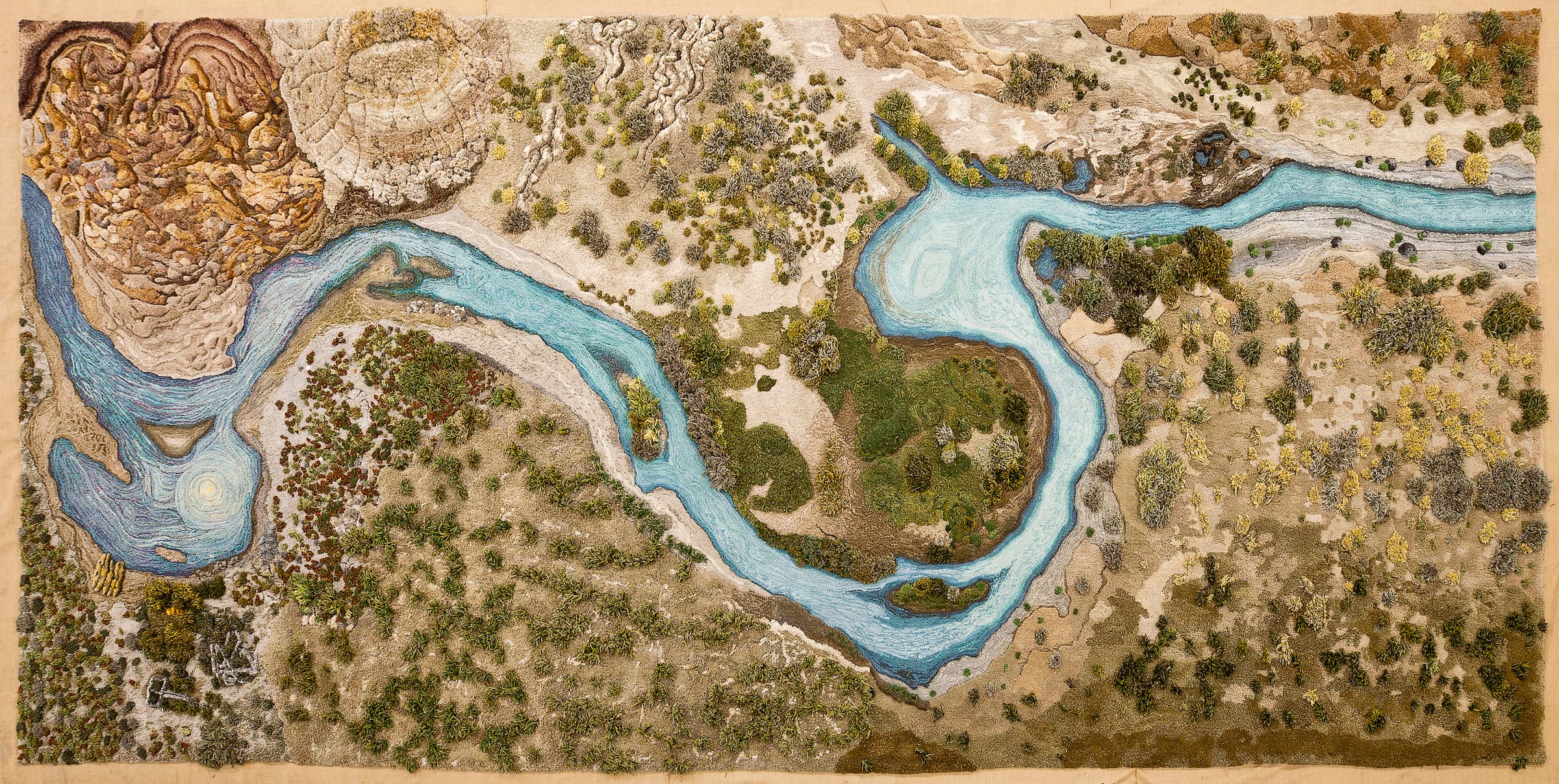 Topographic rug of the Santa Cruz River in Argentina. The light blue river twists and turns through the center of the rug. The surrounding land on the top and bottom is different shades of light browns and dotted with dark greens.