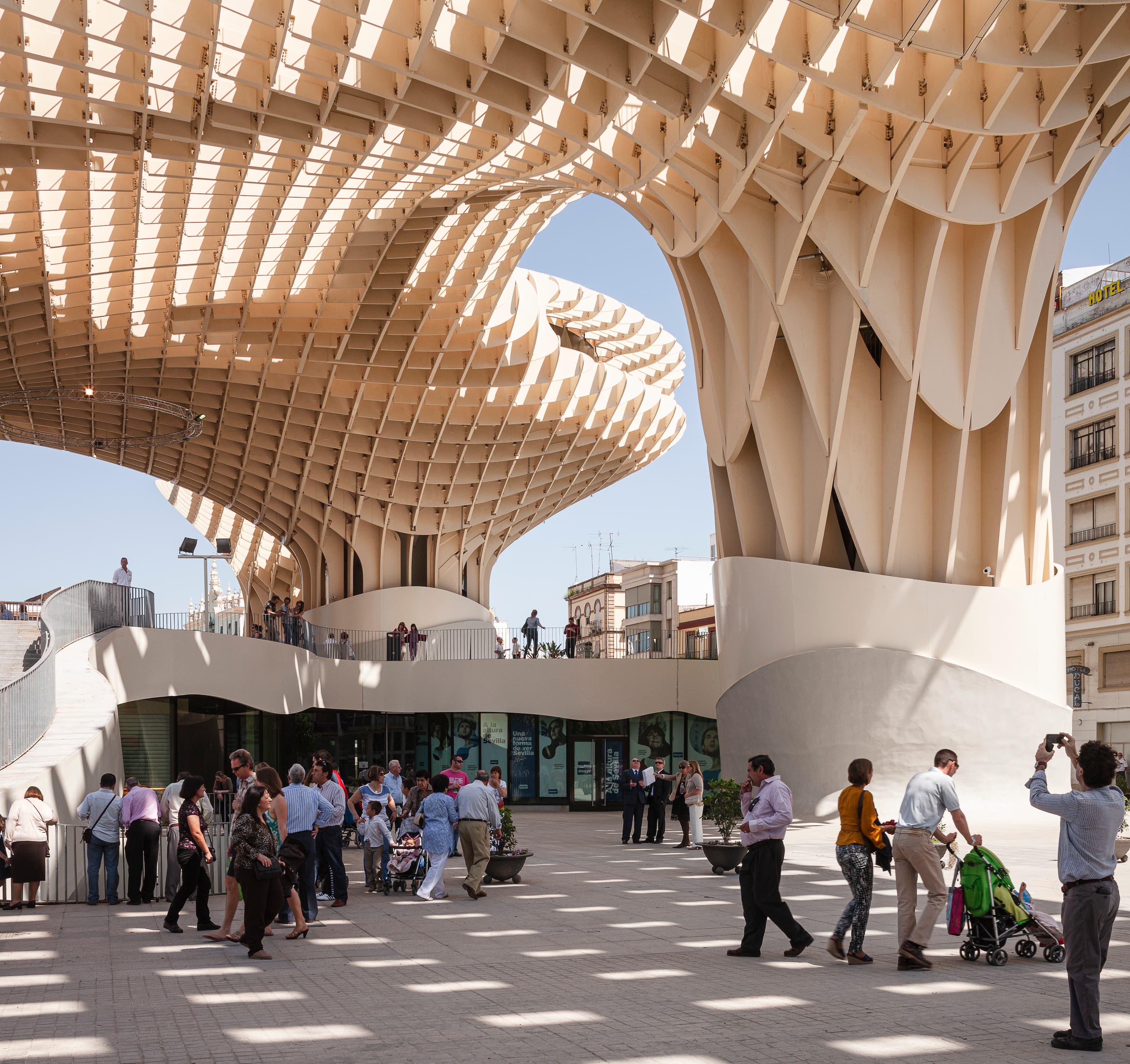 Color photograph of the plaza underneath the Metropol Parasol in Seville, Spain. Light shines through the latticed roof casting a checker-like pattern on the ground. Dozens of people mill about.