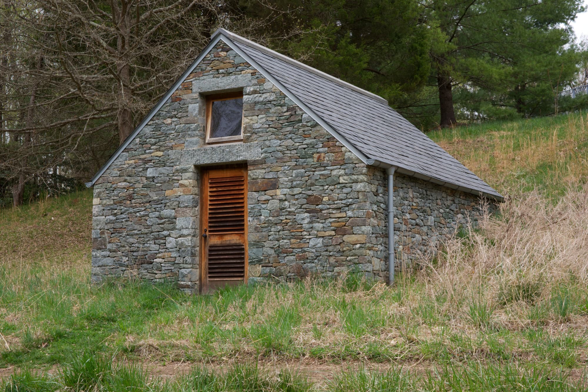 A small stone house built into the side of a grassy green hill. It has a door with a window above it in the center, and a pointy roof. Green trees in the background.