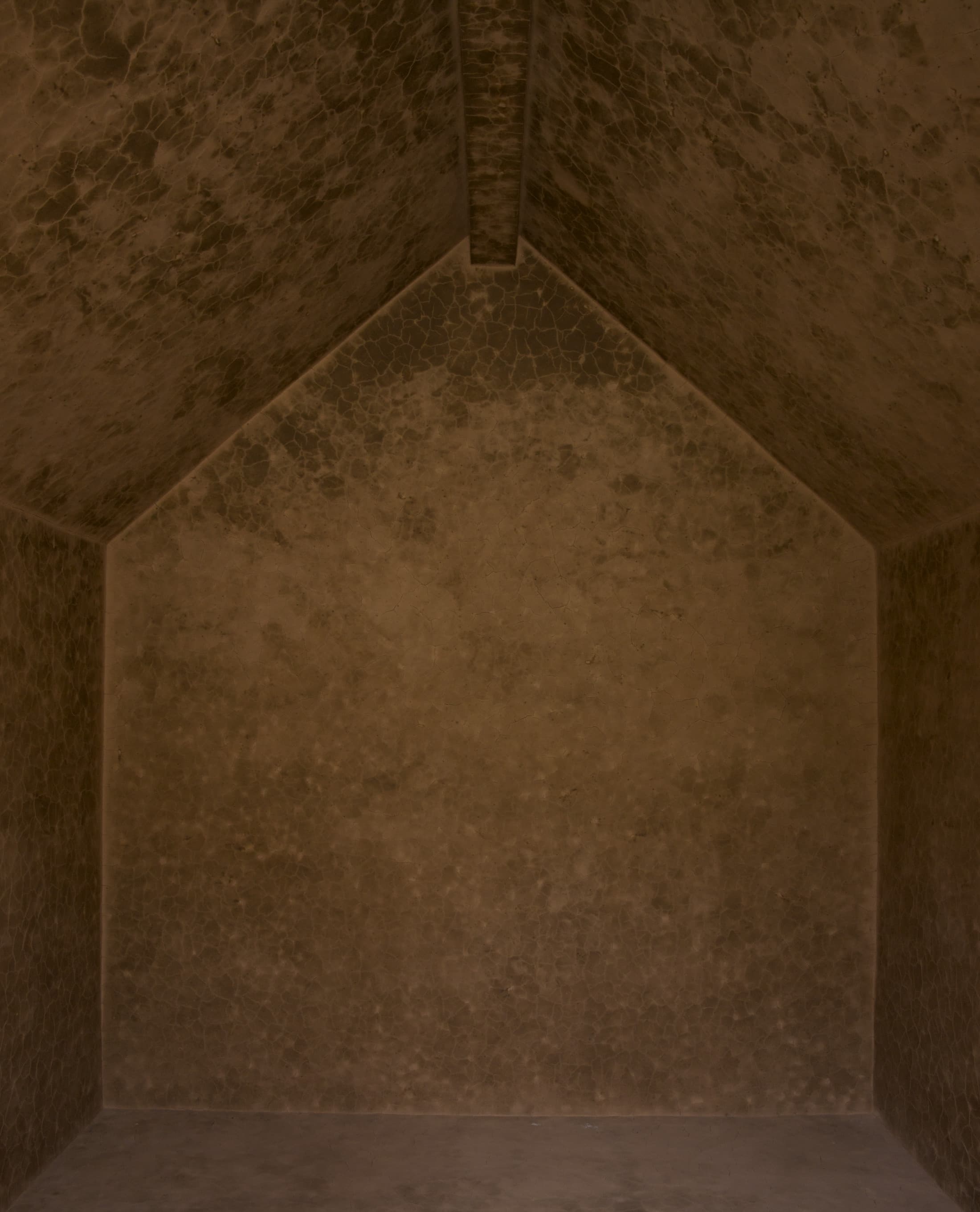 View of interior of *Clay Houses*. Empty room with a lofted ceiling that comes to a point. The light brown walls look like cracked clay.