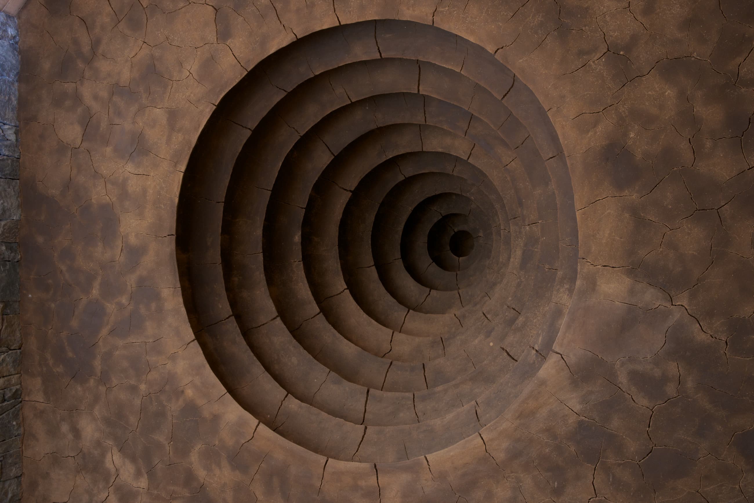 Close-up image of a wall from *Clay Houses* that recedes into a series of concentric circles back into the hill outside of the wall. The surface is varying shades of brown and looks like cracked clay.
