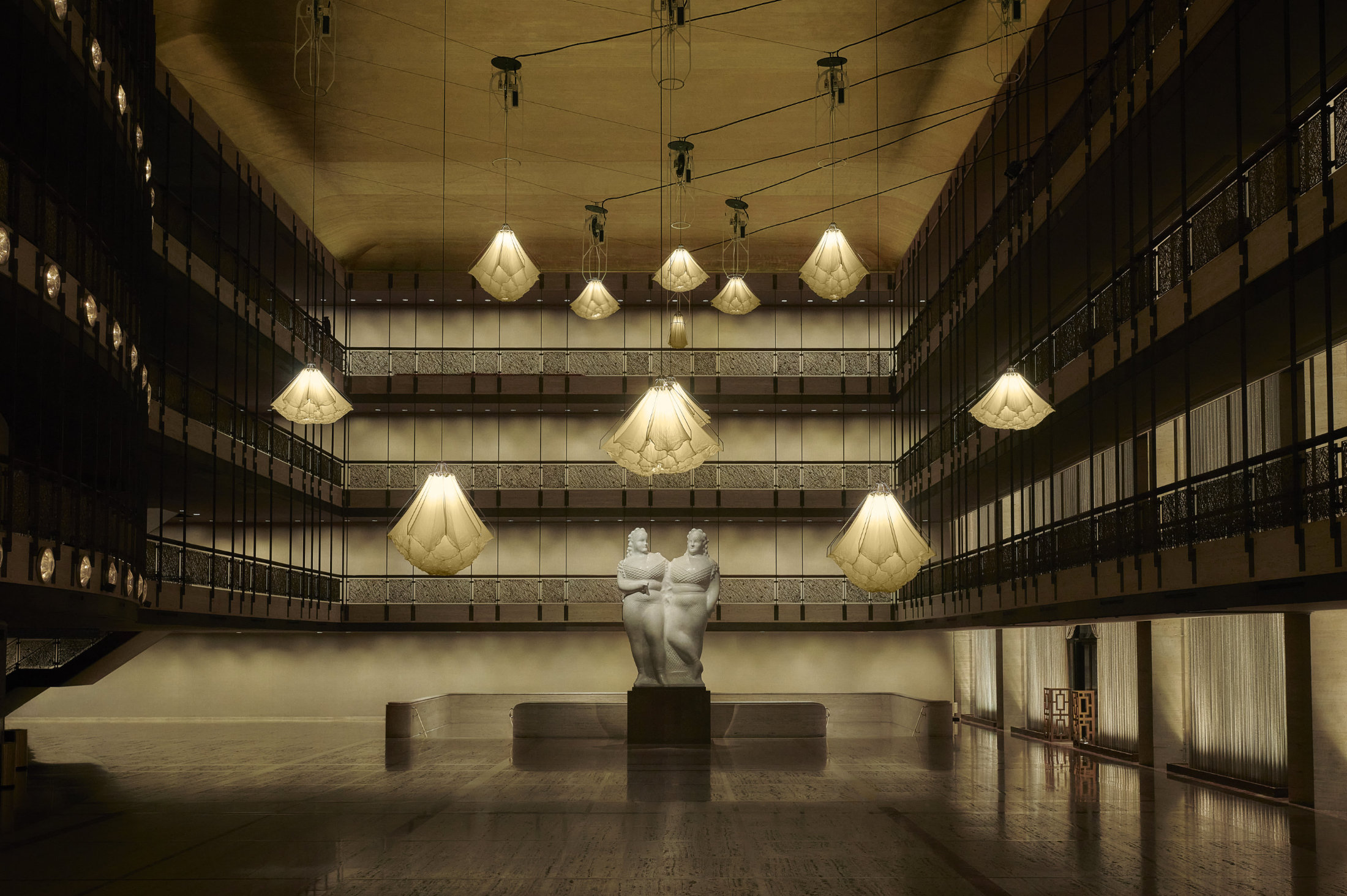 Color photograph of Shylight displayed in the New York City Ballet Theater. Twelve silk chandeliers hang at different levels from a high ceiling. They are all shaped like a flower bloom, but at various stages of opening. A warm, soft light emanates from the chandeliers.