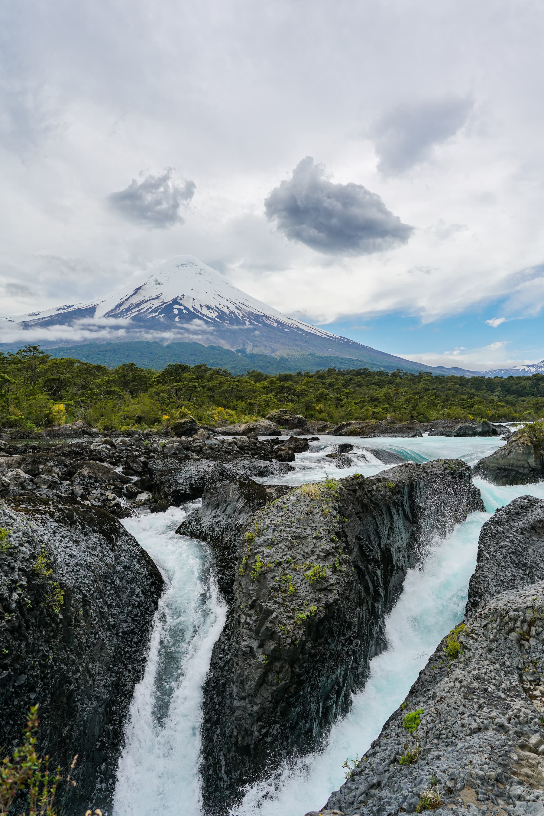 A vertical photograph of the Osorno Volcano in the Chilean Andes. In the background, a large mass of soft clouds above a snow-capped peak. A swath of green forest at the centerline, with two waterfall streams coursing down dark gray rock formations in the foreground.