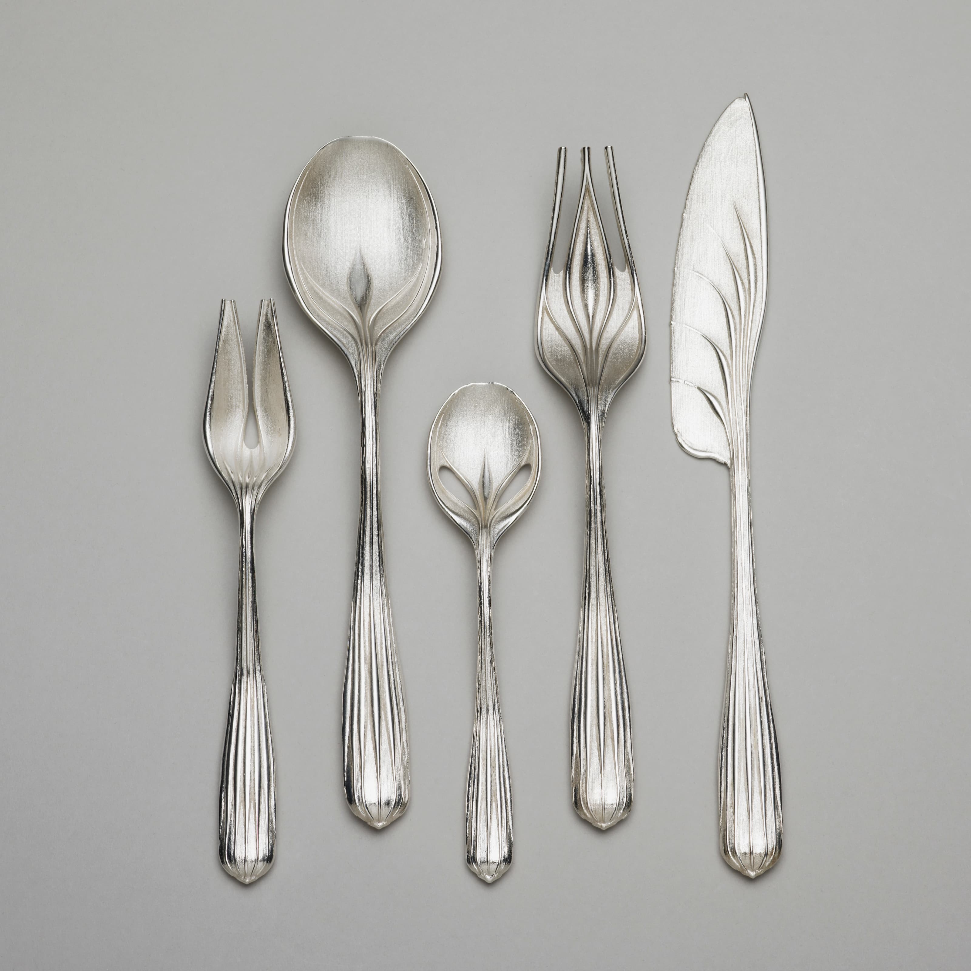 A five-piece, silver, botanical-themed flatware set with two types of forks, two types of spoons, and one knife. The base of each piece is bulbous with vertical lines going up the handle, reminiscent of a seed.
