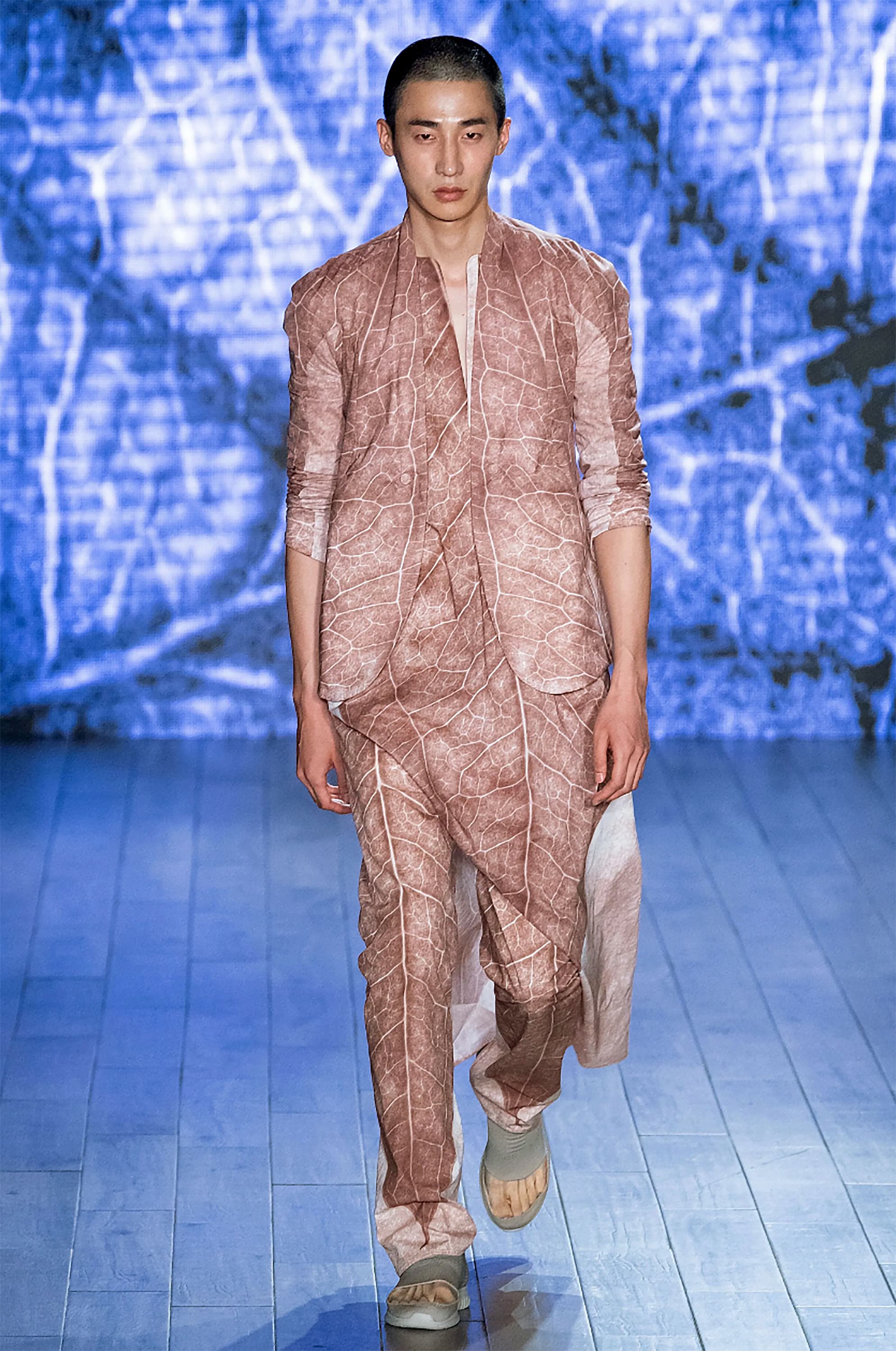 Front view of a model walking down the runway wearing a three-piece suit with a light-brown, autumn leaf print.