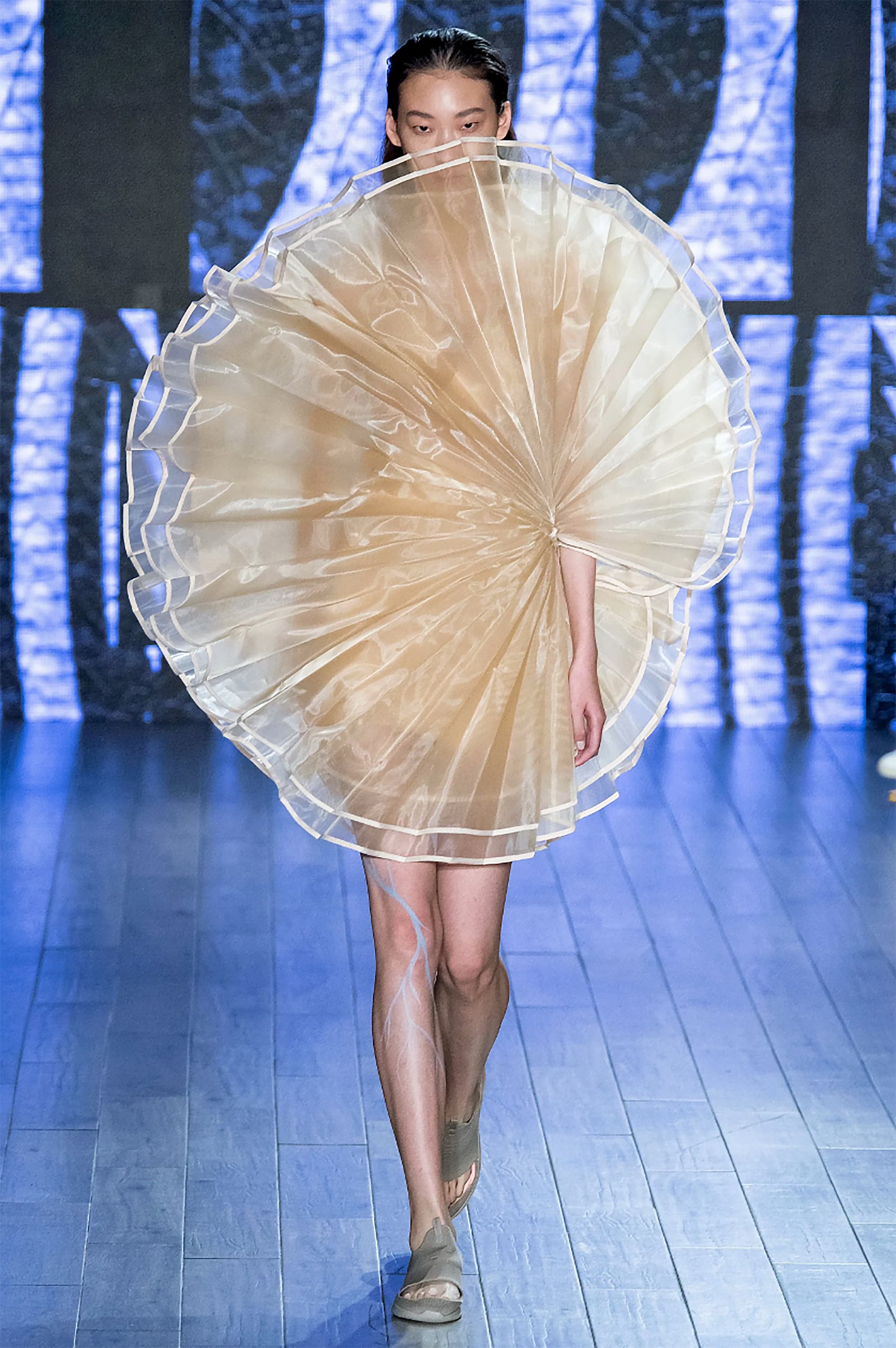Front view of a model walking down the runway wearing a short, ivory colored dress. The dress is circular with the bottom of the circle mid-thigh and the top covering the bottom half of the model’s face. Large pleats radiate around the dress like a folding fan.