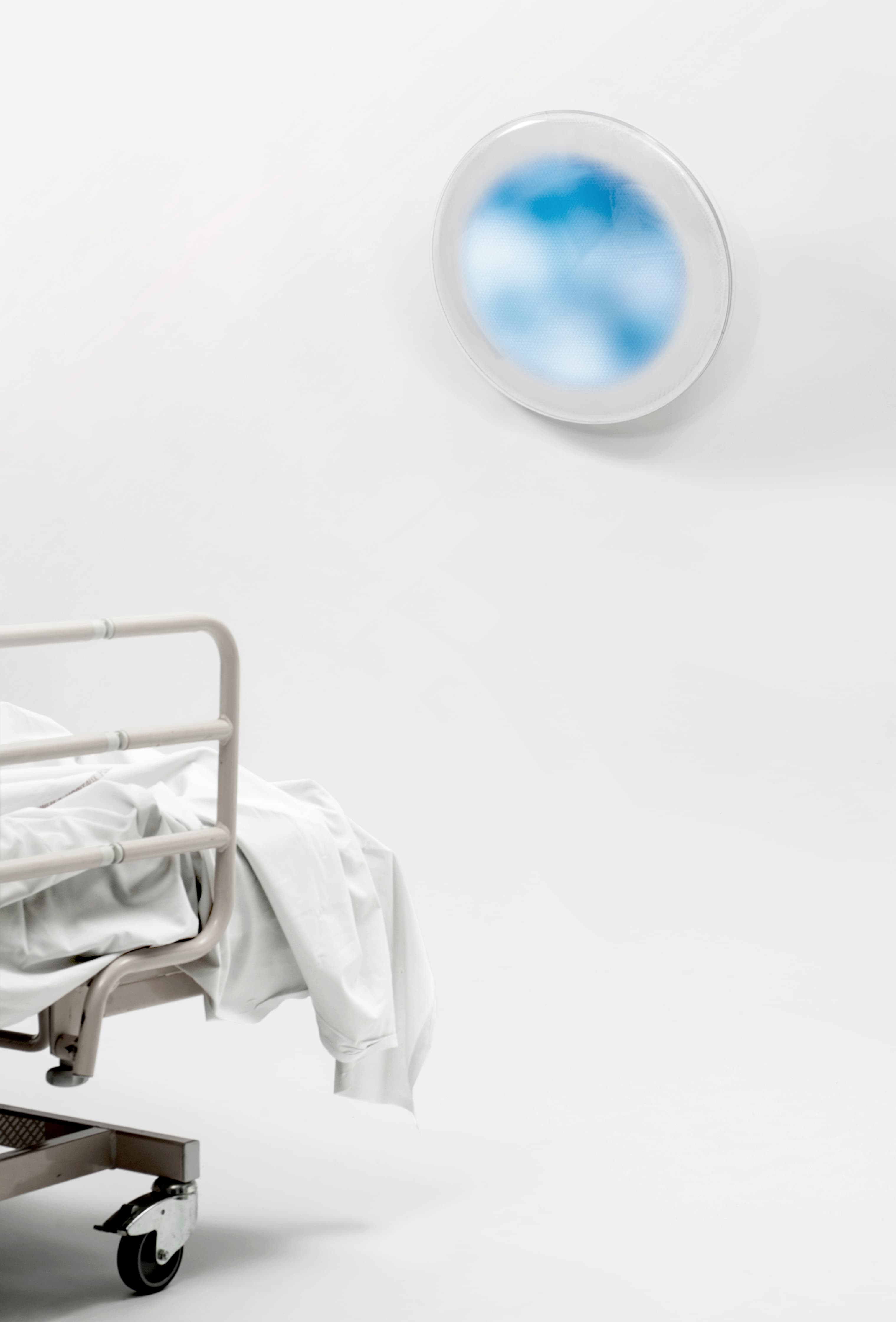 In the bottom-left corner, the bottom edge of a hospital bed with a blanket hanging over the edge. In the top right corner, the artwork that looks like a circular window to a blue sky with white clouds.