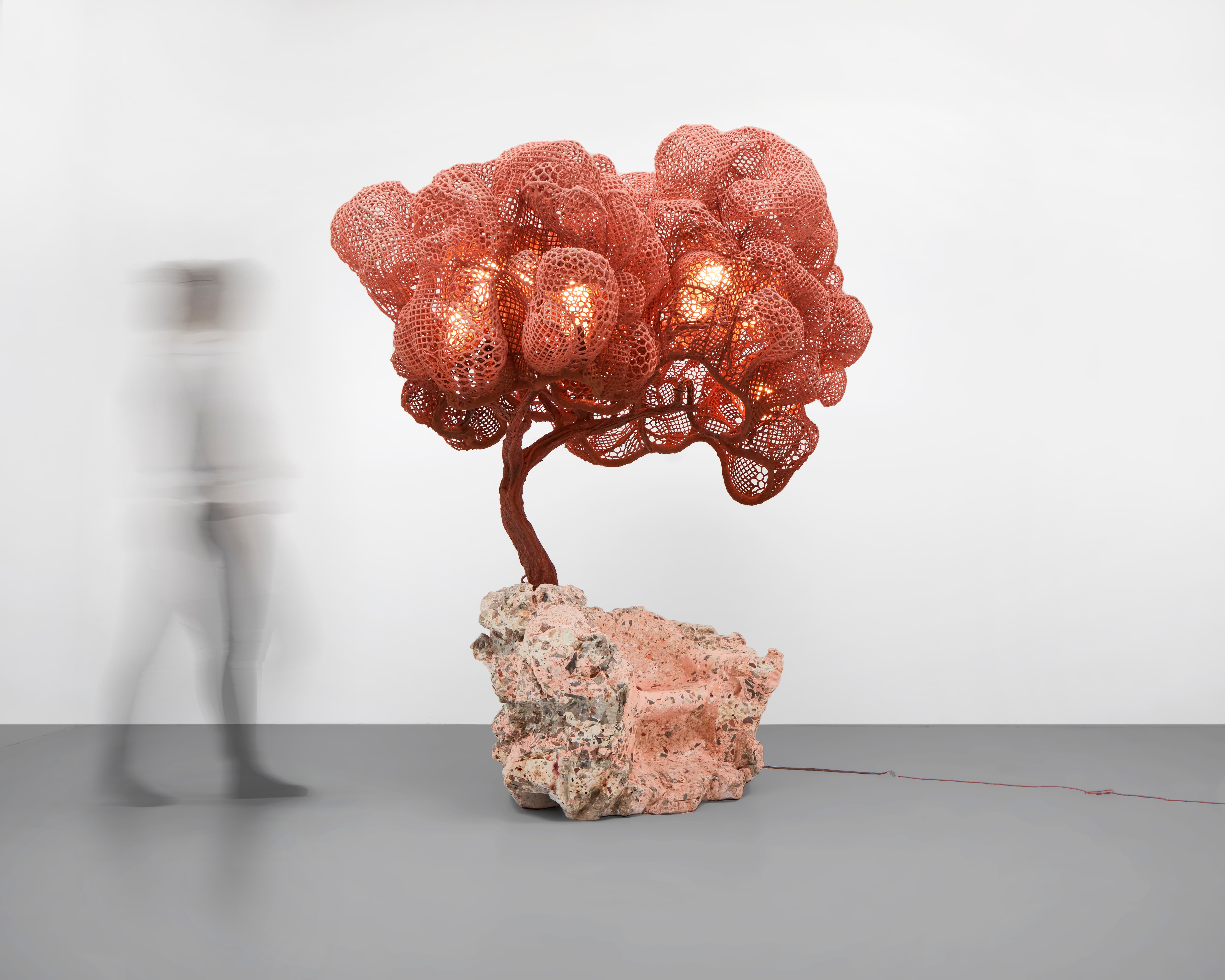 A big armchair, from which one large metal trunk grows with a metal mesh cork presenting as the crown of a tree. The seating area is pink with gray flecks, reminiscent of pink granite. The trunk is a sienna brown, and the canopy of the tree is dusty pink.