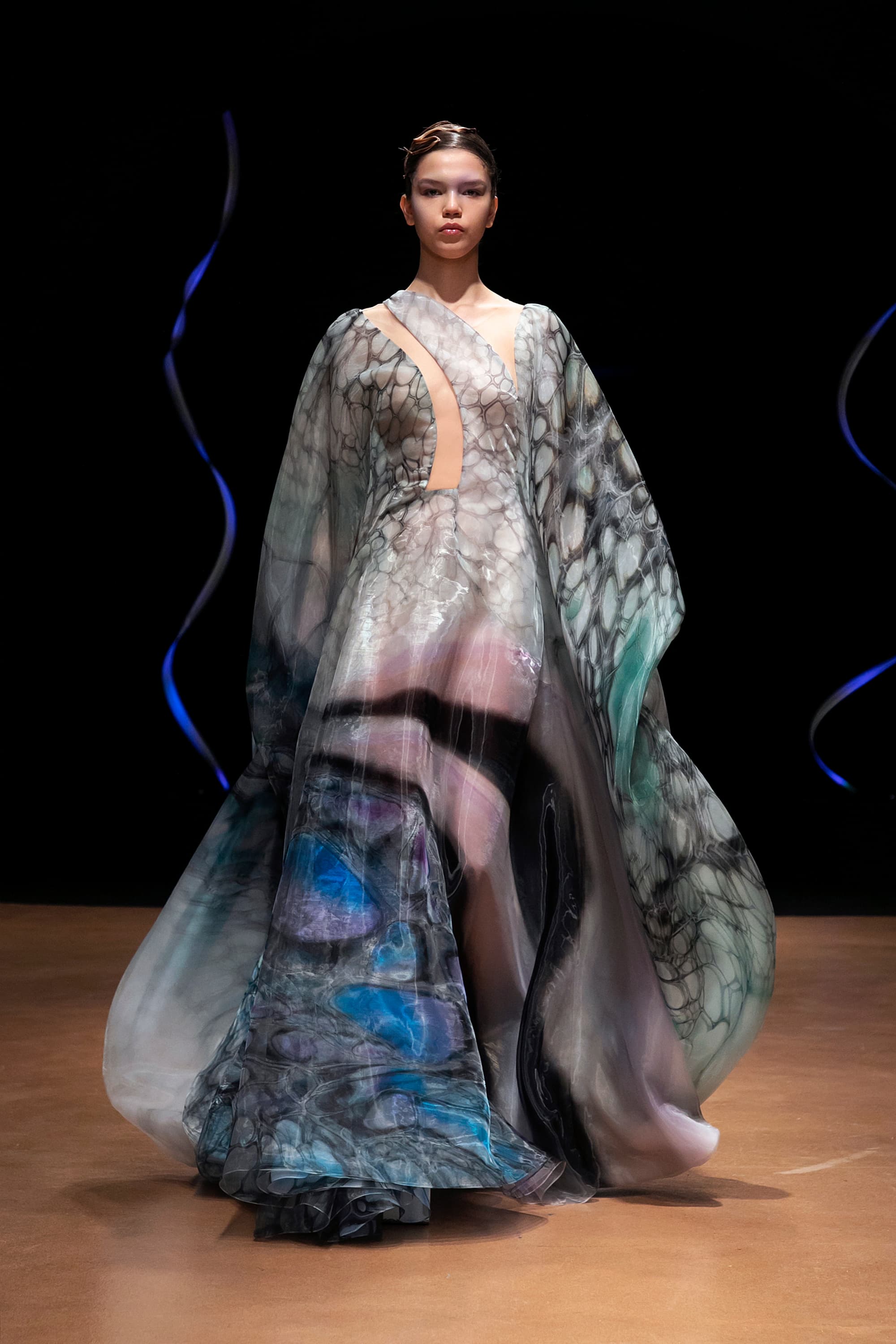 Front view of a model wearing a floor-length dress with long sleeves that completely cover their arms. The drapey material has an aquatic print in seafoam green, black, blue, and purple.