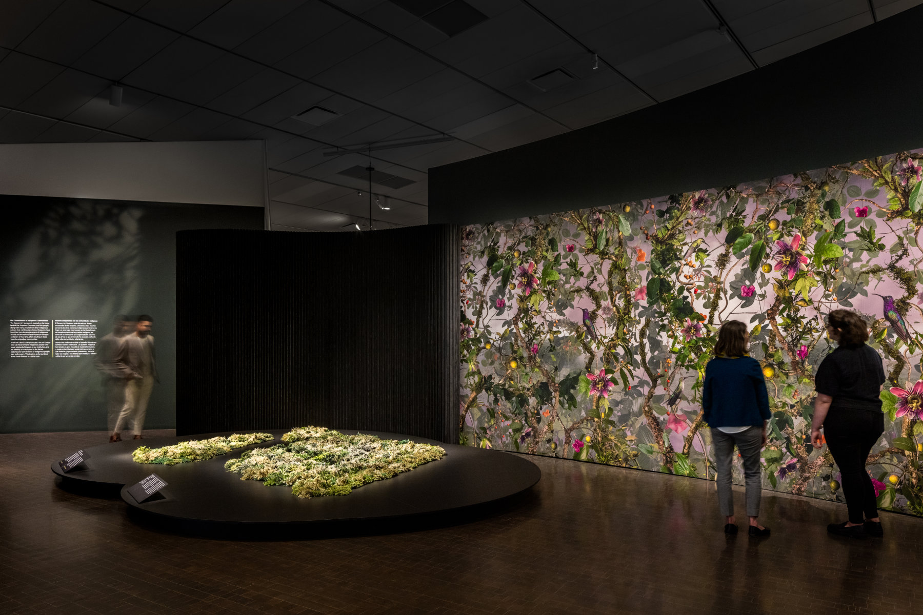 Two people view a large wall with a floral pattern with pinks and greens, and another person views a green rug on a low pedestal.