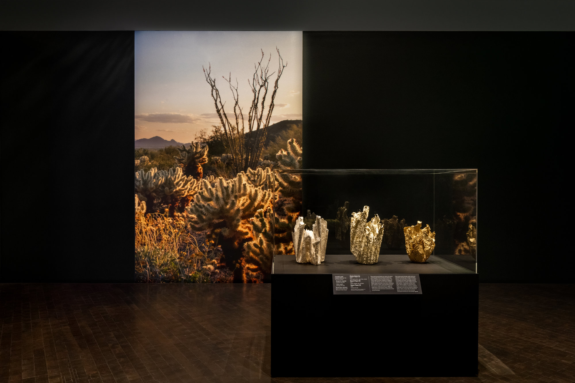 Three textured vessels sit in a display case and match the golden hues of a photograph of the desert on the wall behind.