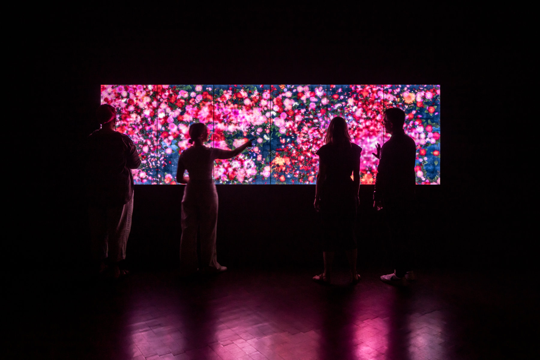Four people view a brightly glowing screen with vibrant pink flowers in a darkened room.