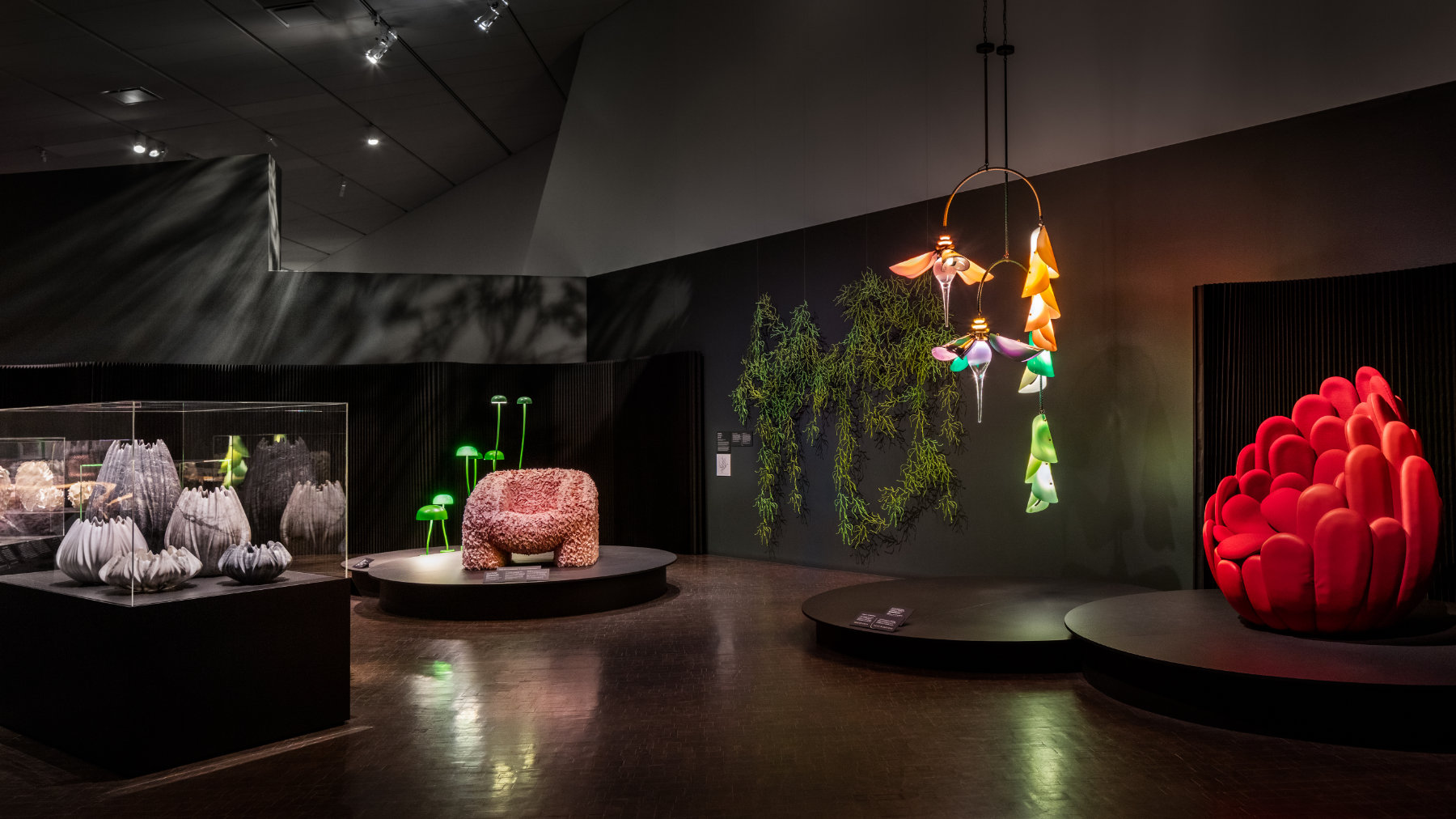 Two chairs, a chandelier, a wall-based sculpture, lamps, and several sculptural vessels are on view in this dark brown gallery.
