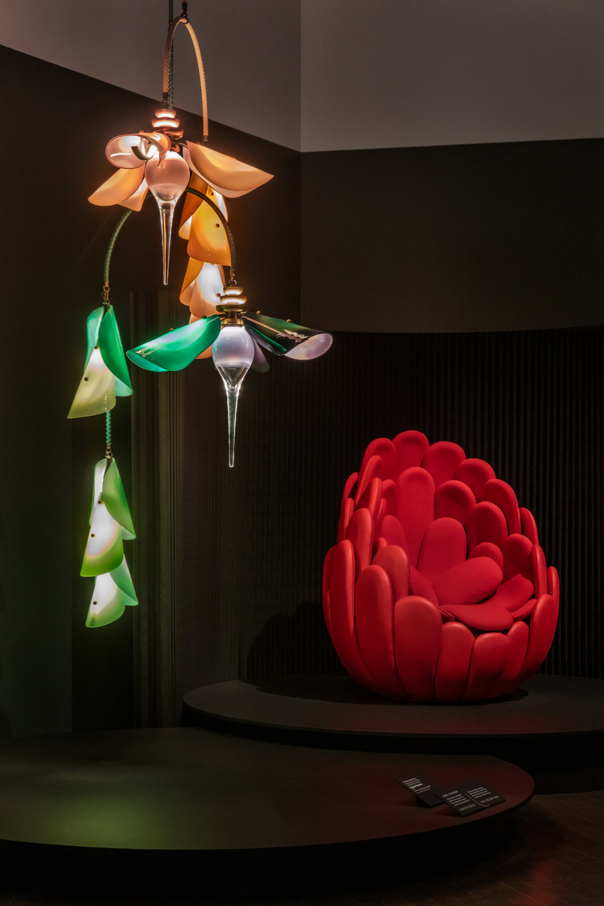 A chandelier that evokes the shapes and colors of hummingbirds hangs near a bright red chair in a brown gallery.
