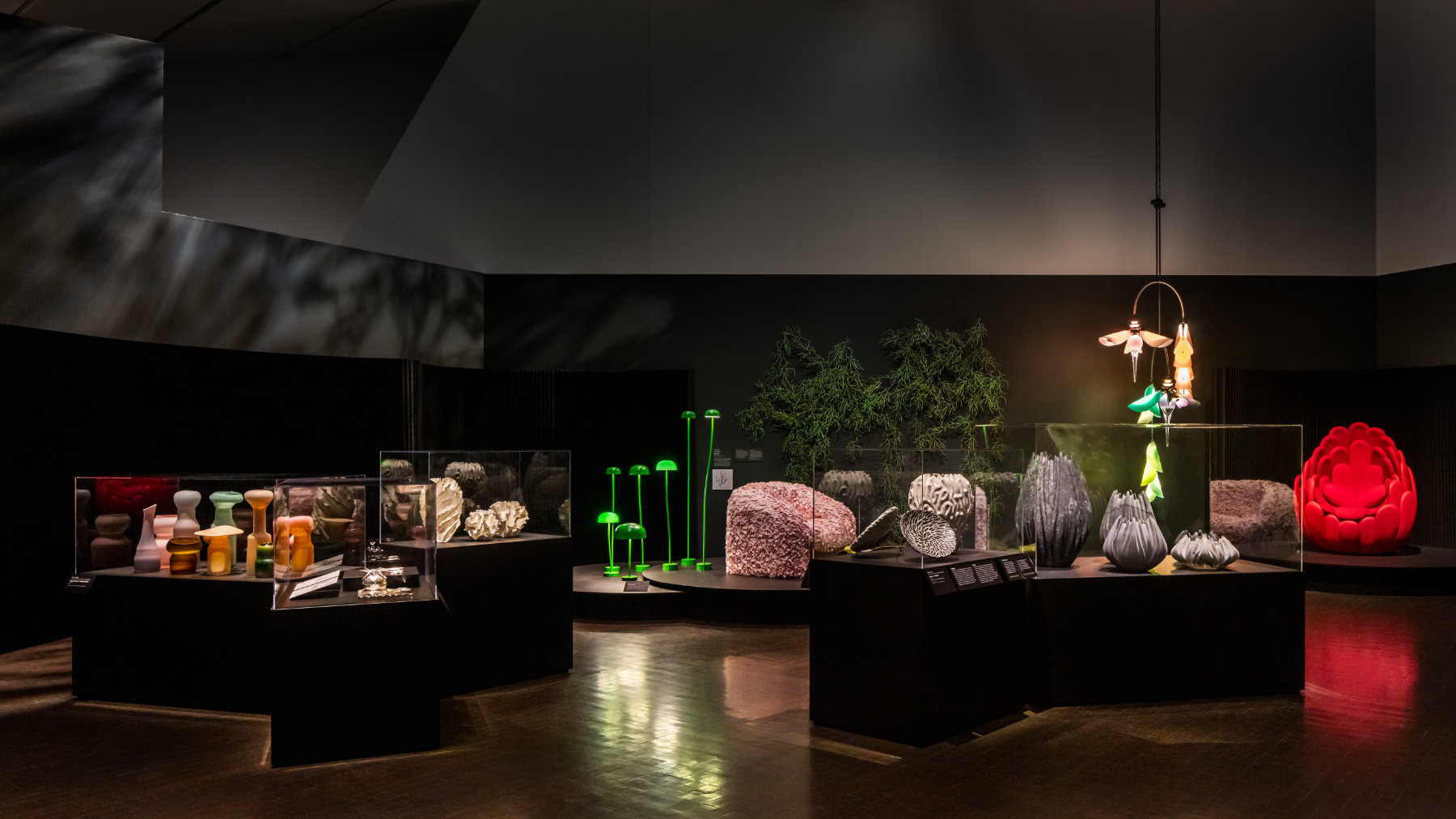 Five display cases feature sculptural vessels and cutlery in front of two chairs, a chandelier, a wall-based sculpture, and several lamps.