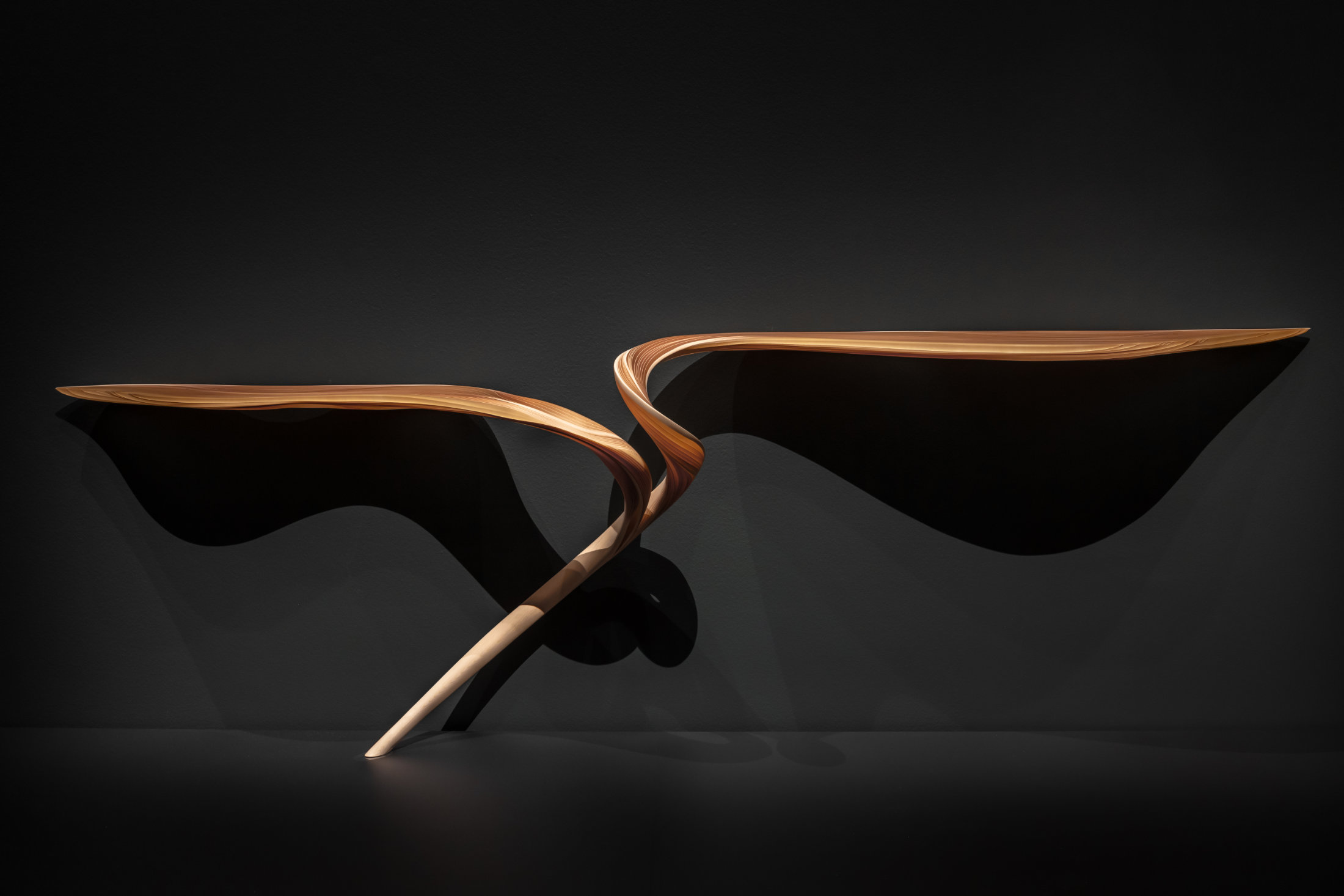 A thin table that evokes a bird in flight rests on one leg against a dark wall.