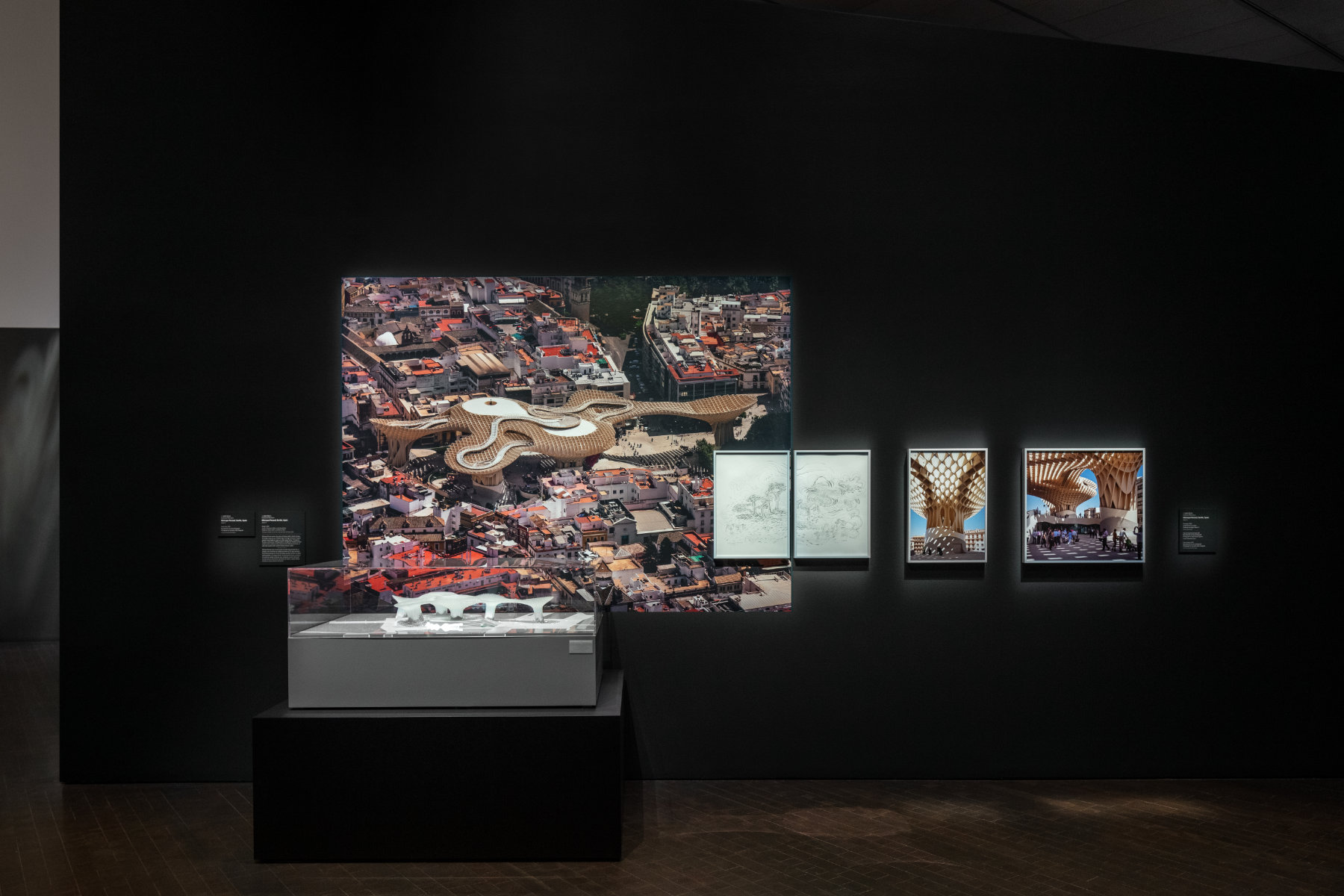 A white architectural model sits in a display case in front of a colorful photograph of the actual building. Four framed drawings and photographs also hang on the dark wall.
