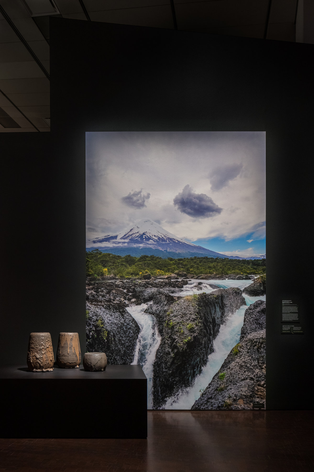 Three dark vessels sit on a pedestal in front of a large image of a mountain in the background and a river flowing through rocks in the foreground.