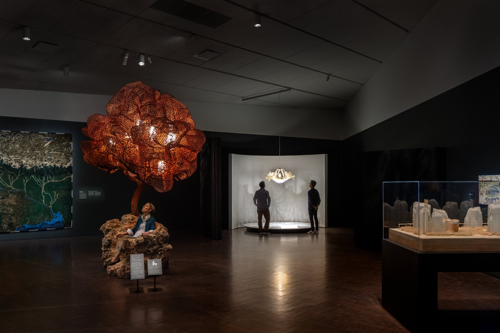In a dark gallery, two people view a lit chandelier, one person sits on a craggy seat under a lit tree-like form, an image of a landscape hangs on one wall, and an architectural model sits in a display case.