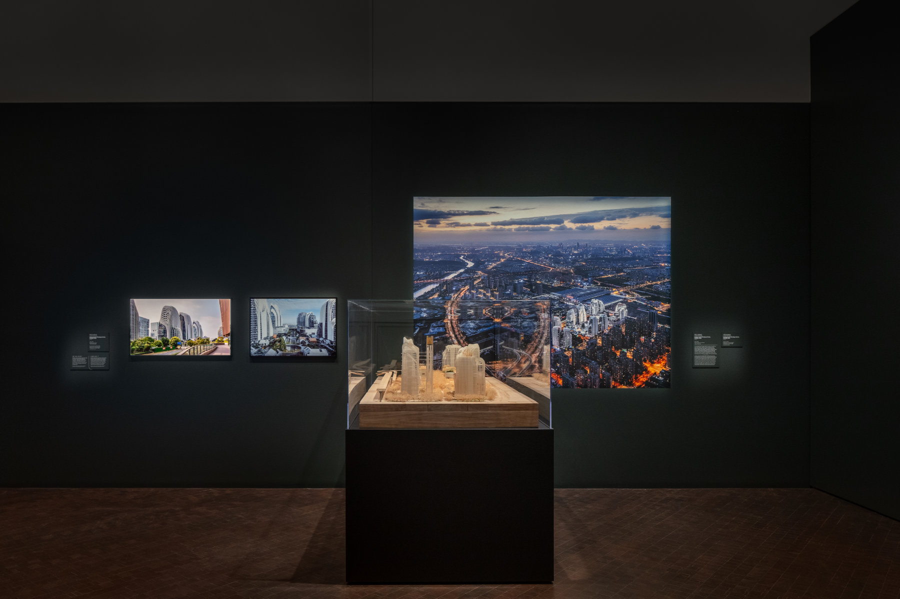 A pale architectural model sits in a display case in front of a large colorful photograph of the actual building. Two framed photographs of the building also hang on the dark wall.