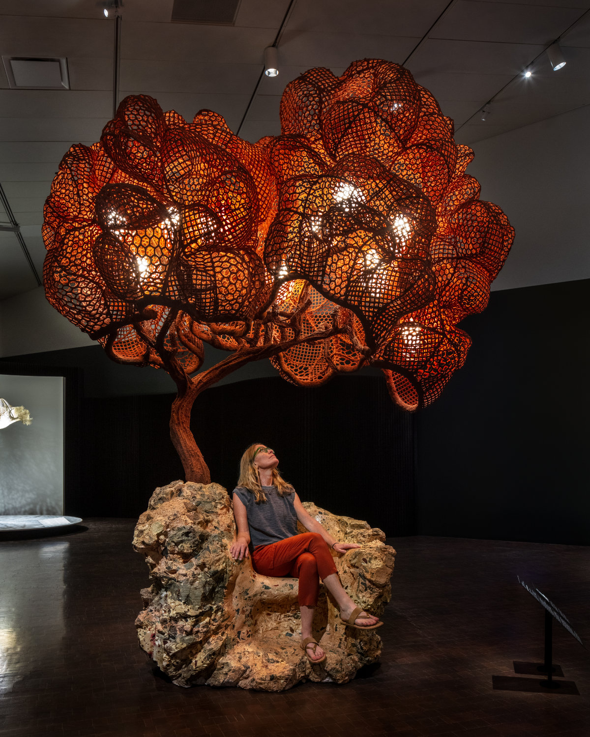 A person sits on a seat that looks like a rock outcropping and looks up at the form of a copper-colored form that evokes a tree, which is lit from the inside.