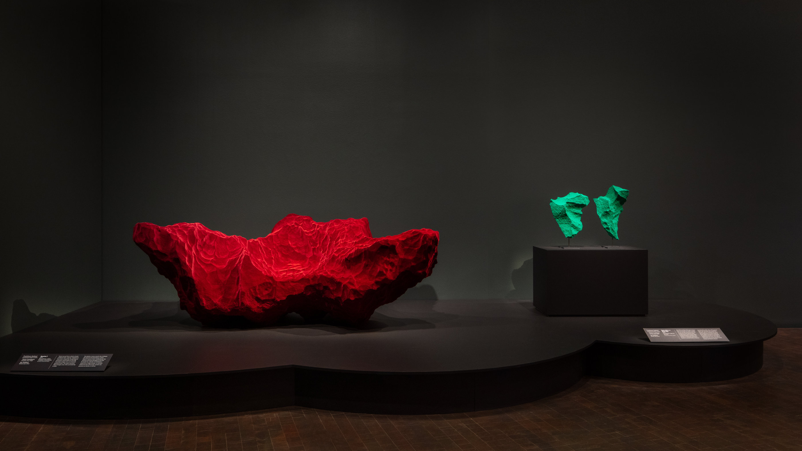 A deep-red sofa that evokes a rock formation sits on a low pedestal next to two bright green forms on a taller pedestal that also evoke rocky formations.