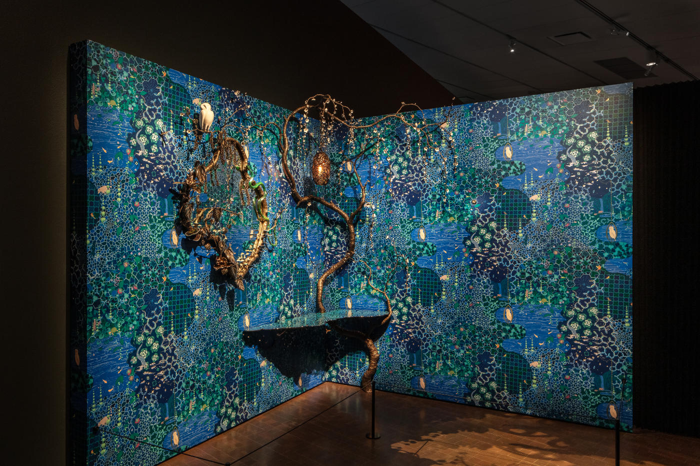 Predominantly blue wallpaper adorns two walls that meet in a corner. The frame of the mirror on the left wall evokes tree branches, and a copper-colored tree-like sculpture extends the full height of the installation.