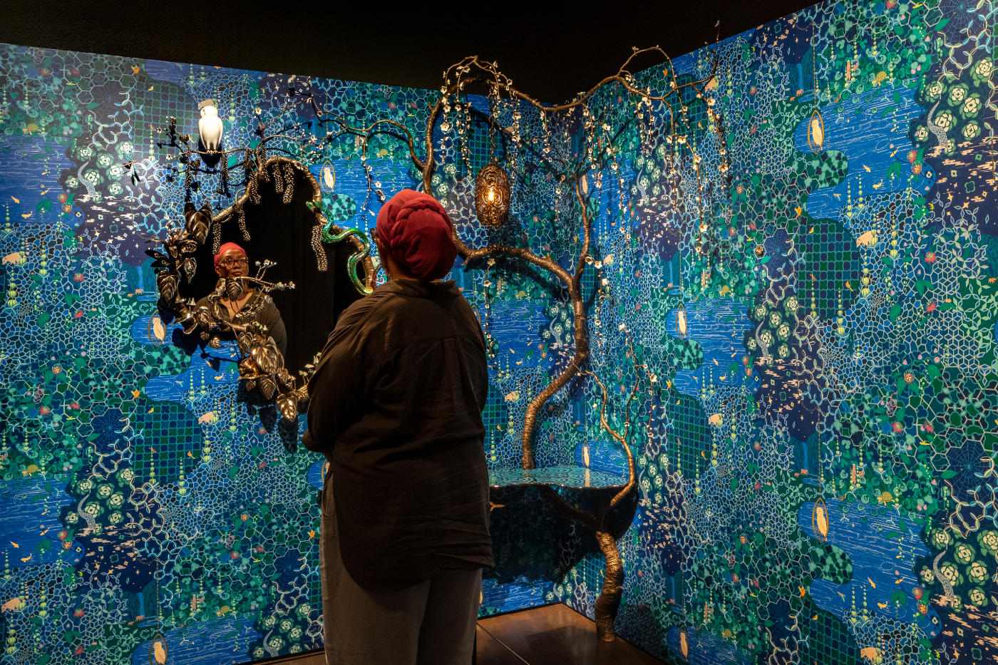A person views themselves in the mirror on the left wall of Midnight in the Meadow. Predominantly blue wallpaper adorns two walls that meet in a corner. The mirror's frame and a tall sculpture evoke organic tree branches.