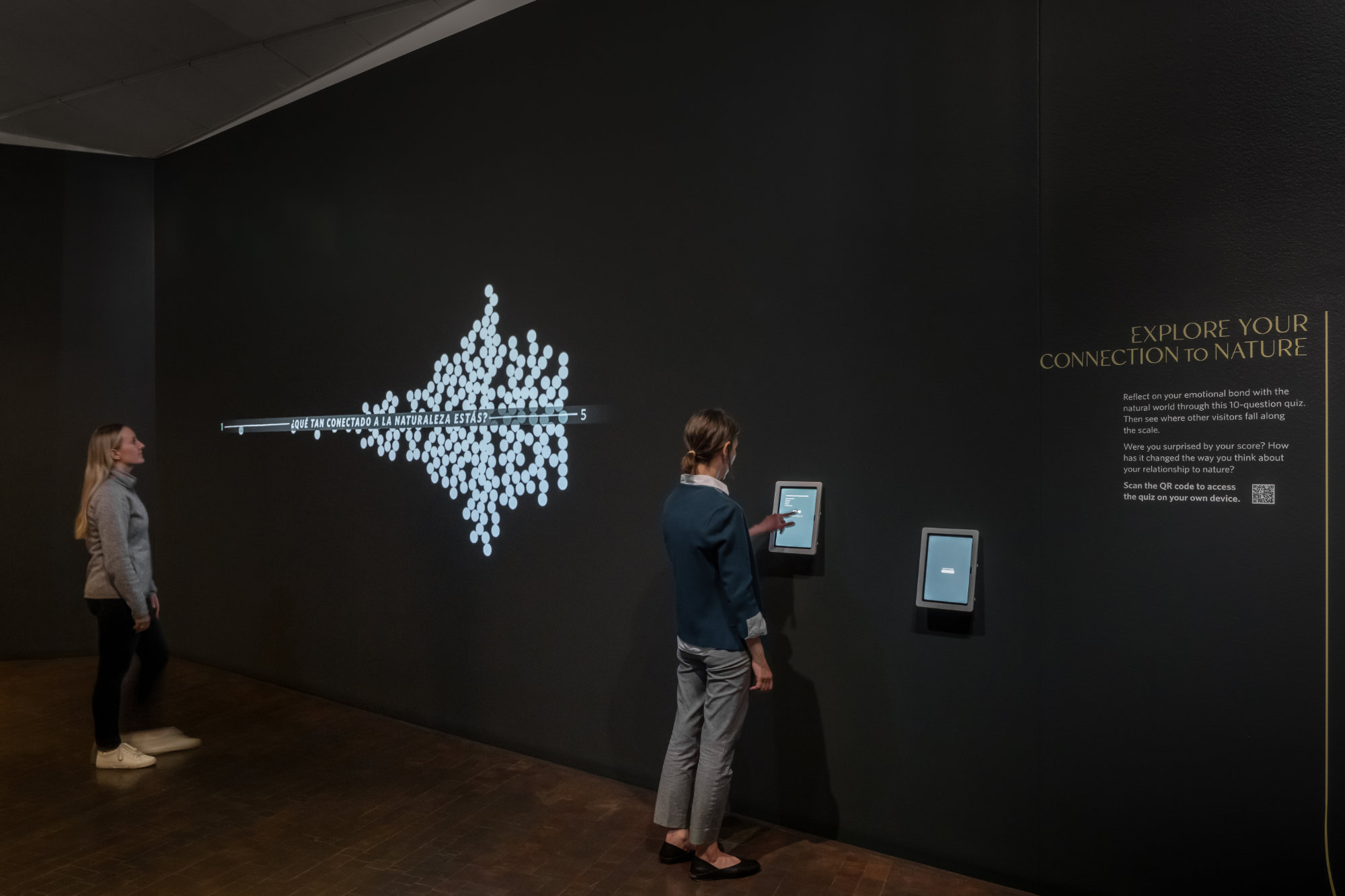 In a dark gallery, one person views a projection of grouped dots on the wall with the phrase Que Tan Conectado a la Naturaleza Estas? Another person interacts with a digital screen on the wall. Light-colored text  reads Explore Your Connection to Nature