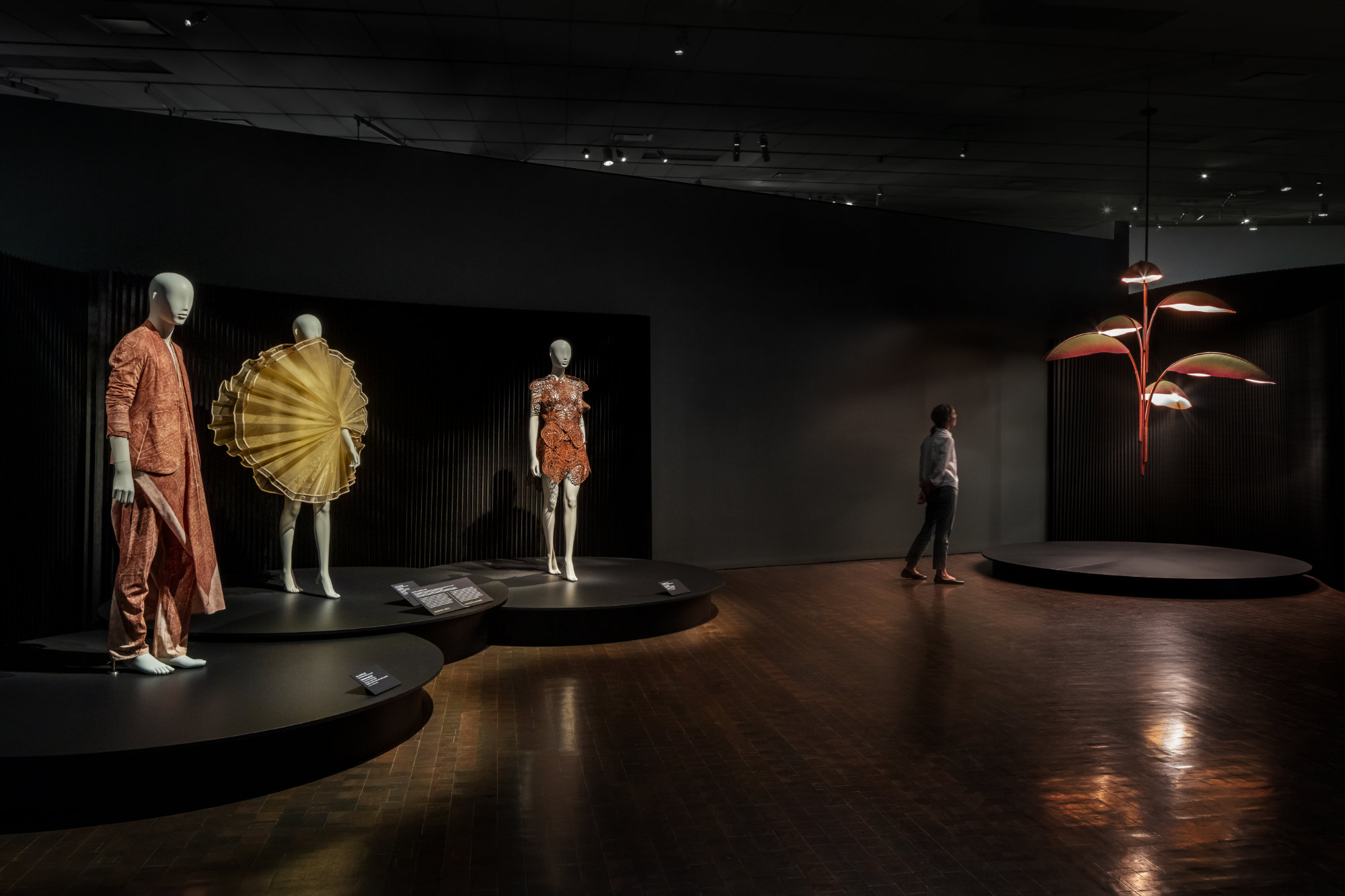 Three white mannequins stand on a low pedestal in a dark gallery. One wears a reddish-brown tunic suit with the pattern of leaf veins. One wears a circular yellow gown that extends from nose to knees. The third wears a short reddish-brown dress with fabric laser cut in the shape of leaves. A person views a chandelier that evokes large drooping leaves on the right.
