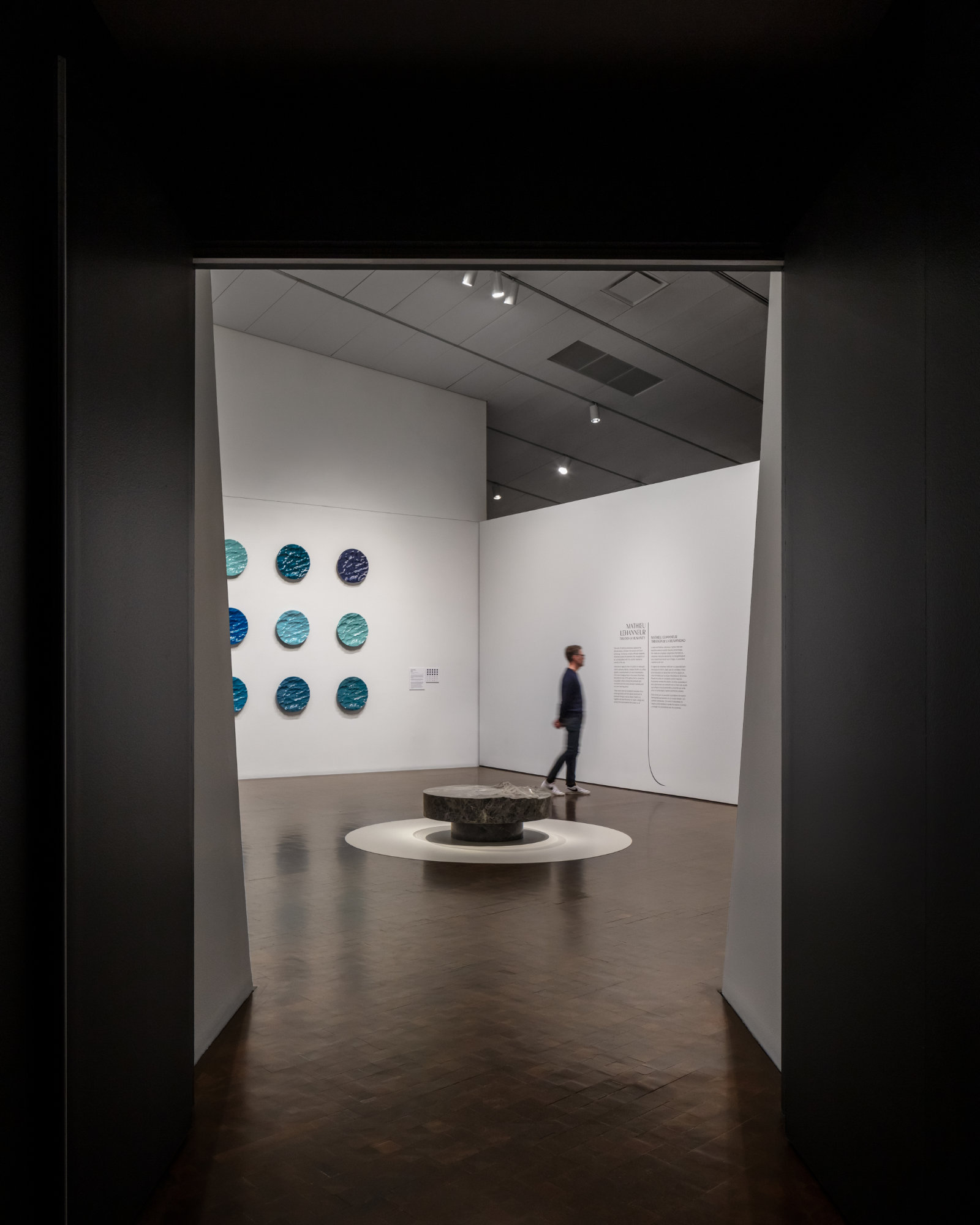 A dark doorway frames a brightly lit gallery. A person reads text on the wall in front of a low sculpture that evokes waves captured in stone. On the left wall hang circular forms in a variety of blue hues.
