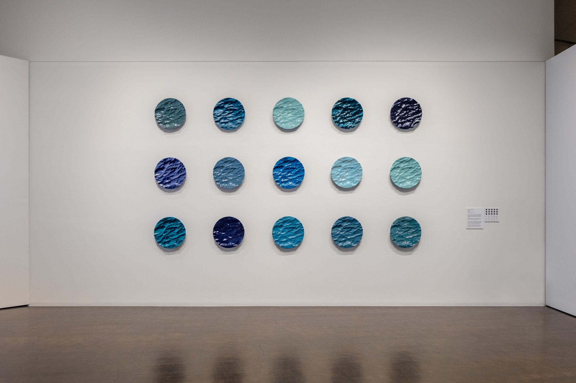 A grid of 15 circular forms hangs on a white wall. They evoke waves captured in sculptural form and feature a variety of blue hues.