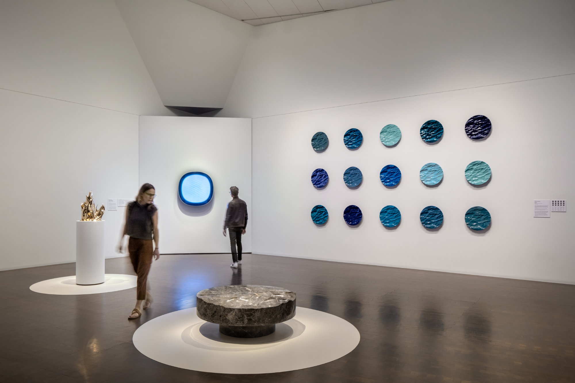 Two people view art in a white gallery. A low stone sculpture in the center and a group of 15 wall-based blue sculptures evoke waves captured in stone. A gold sculpture on the left evokes flames. A screen on the rear wall exudes pale blue light.